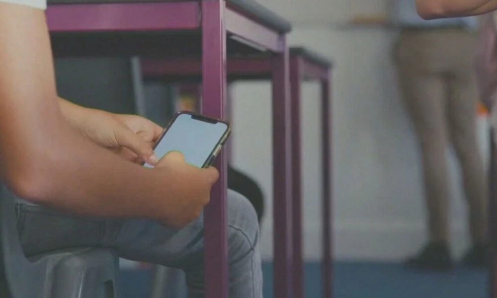 Person seated, using a smartphone; purple chair frames are visible in the background (indoor setting).