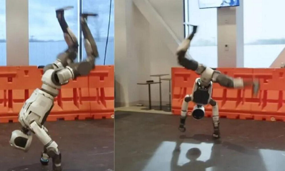 Two humanoid robots performing backflips on a lab floor with orange safety barriers in the background, mid-air with arms raised.