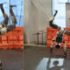 Two humanoid robots performing backflips on a lab floor with orange safety barriers in the background, mid-air with arms raised.