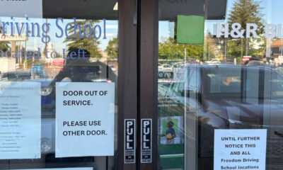 Glass storefront door with multiple signs; main message reads 'DOOR OUT OF SERVICE. PLEASE USE OTHER DOOR.' and 'Driving School' window text visible.