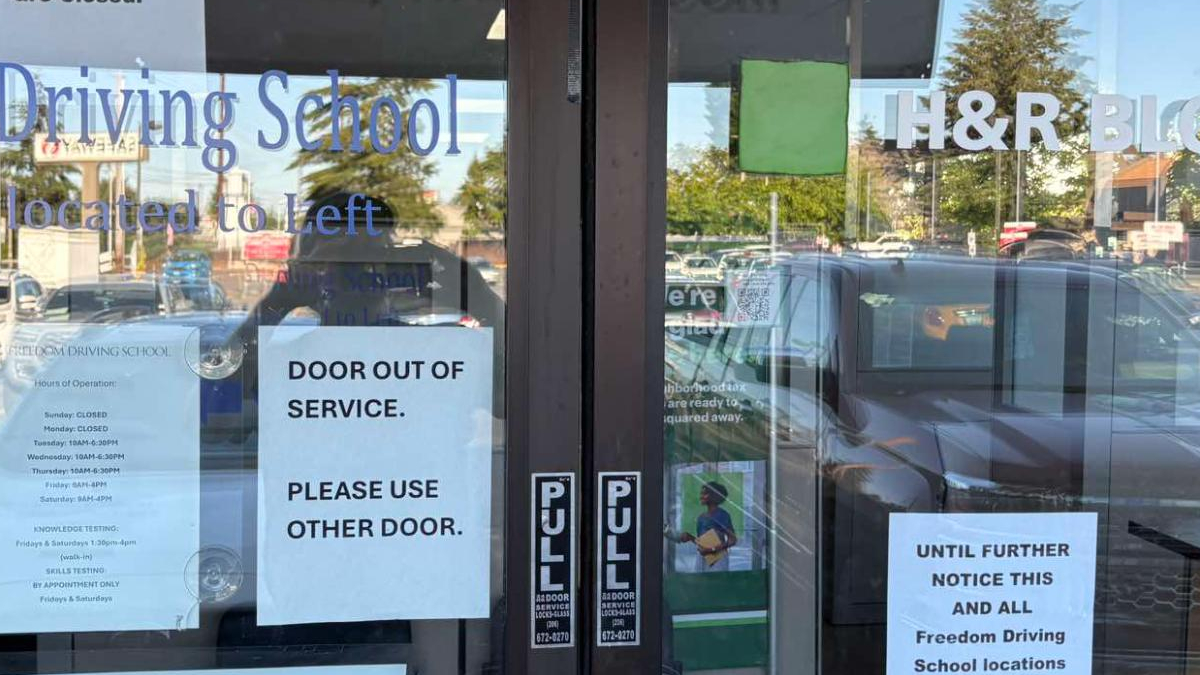 Glass storefront door with multiple signs; main message reads 'DOOR OUT OF SERVICE. PLEASE USE OTHER DOOR.' and 'Driving School' window text visible.