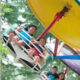 Group of people riding a circular swing ride with a large yellow disk overhead, outdoors among trees.
