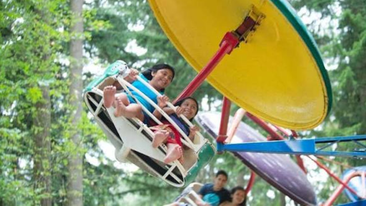 Group of people riding a circular swing ride with a large yellow disk overhead, outdoors among trees.