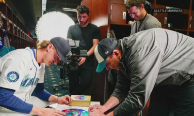 Two Seattle Mariners players sign colorful circular artworks on a table in the locker room as a film crew records them.