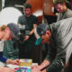 Two Seattle Mariners players sign colorful circular artworks on a table in the locker room as a film crew records them.