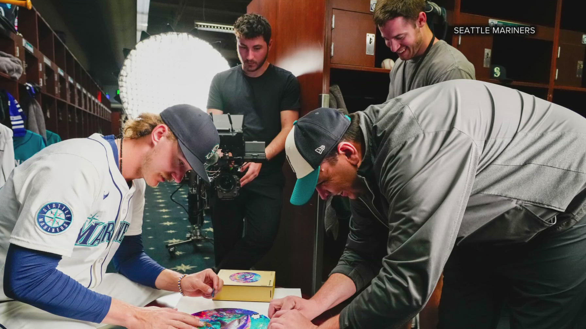 Two Seattle Mariners players sign colorful circular artworks on a table in the locker room as a film crew records them.