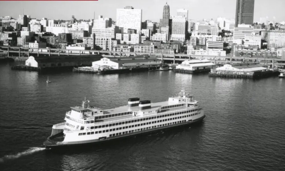 Black-and-white photograph of a large passenger ship cruising in a harbor with a city skyline in the background, showing multiple decks and two smokestacks.
