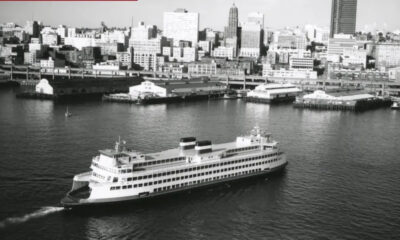 Black-and-white photograph of a large passenger ship cruising in a harbor with a city skyline in the background, showing multiple decks and two smokestacks.