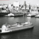 Black-and-white photograph of a large passenger ship cruising in a harbor with a city skyline in the background, showing multiple decks and two smokestacks.