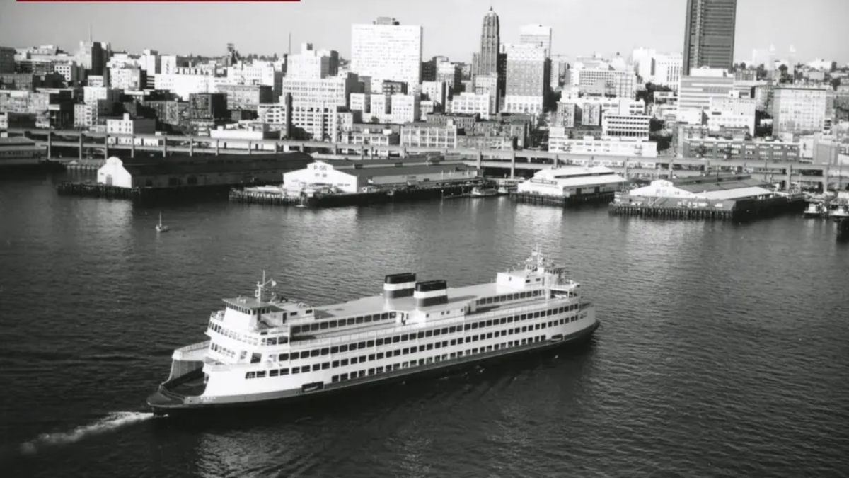 Black-and-white photograph of a large passenger ship cruising in a harbor with a city skyline in the background, showing multiple decks and two smokestacks.