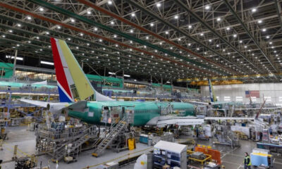 Inside a large aircraft hangar, a teal fuselage jet is on a raised platform surrounded by scaffolding and workers.
