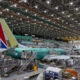 Inside a large aircraft hangar, a teal fuselage jet is on a raised platform surrounded by scaffolding and workers.