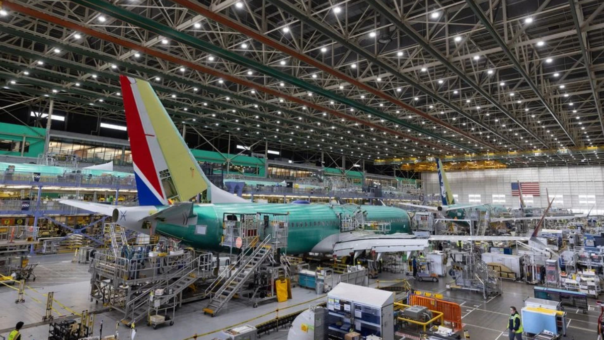 Inside a large aircraft hangar, a teal fuselage jet is on a raised platform surrounded by scaffolding and workers.