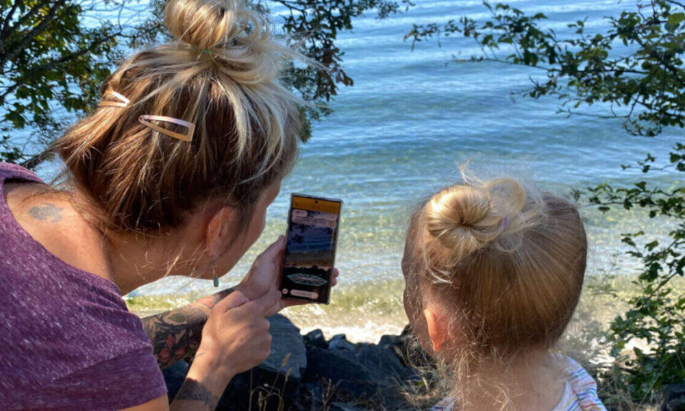 Adult woman and a young girl with blonde bun hairstyles sit by a rocky lakeshore, looking at a smartphone together by the water.