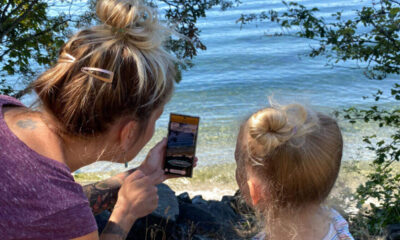 Adult woman and a young girl with blonde bun hairstyles sit by a rocky lakeshore, looking at a smartphone together by the water.