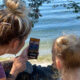 Adult woman and a young girl with blonde bun hairstyles sit by a rocky lakeshore, looking at a smartphone together by the water.