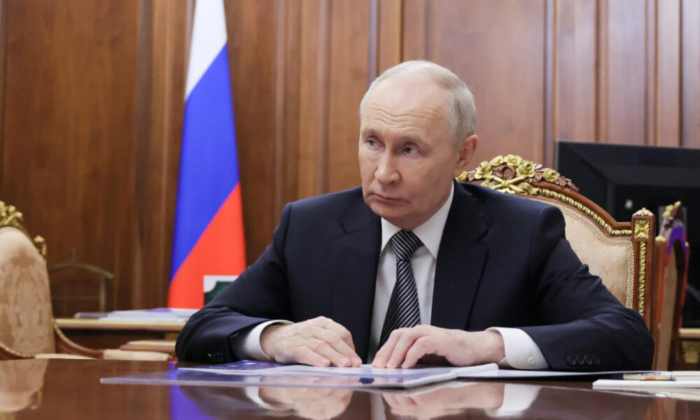 A government official in a dark suit sits at a polished desk with papers, in a wood-paneled room with a flag in the background.