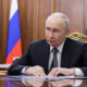 A government official in a dark suit sits at a polished desk with papers, in a wood-paneled room with a flag in the background.