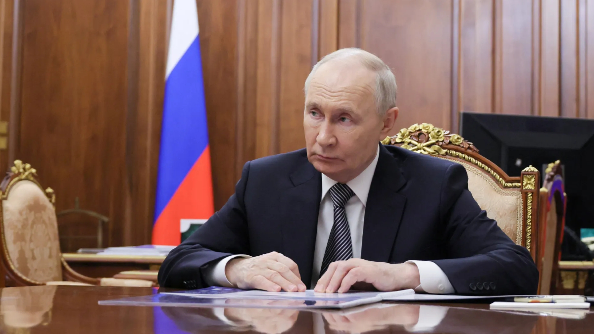 A government official in a dark suit sits at a polished desk with papers, in a wood-paneled room with a flag in the background.