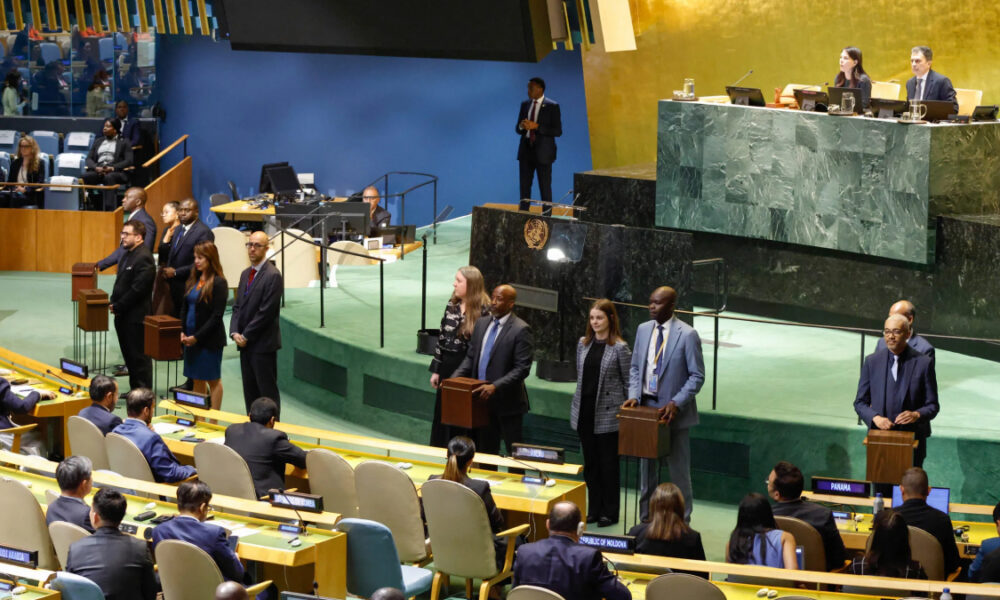 Delegates stand at podiums in the United Nations General Assembly chamber during a session.