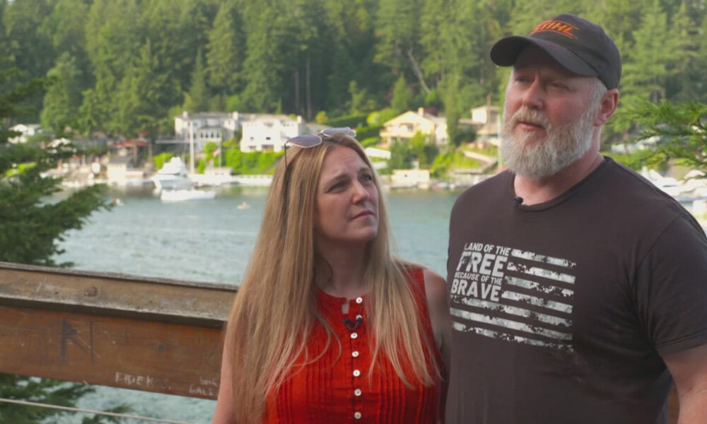 Couple posing by a lakeside railing: a bearded man in a black cap and t-shirt stands beside a woman with long blonde hair in a red sleeveless top, with boats, houses and trees in the background.