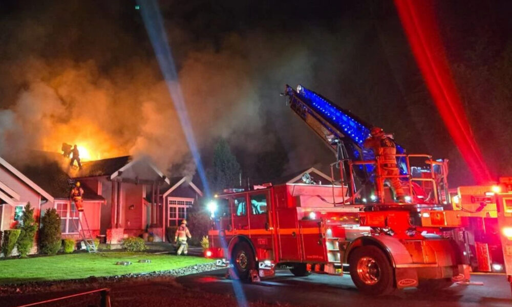 Nighttime fire at a house; flames on the roof, smoke, and firefighters on a ladder battling the blaze.
