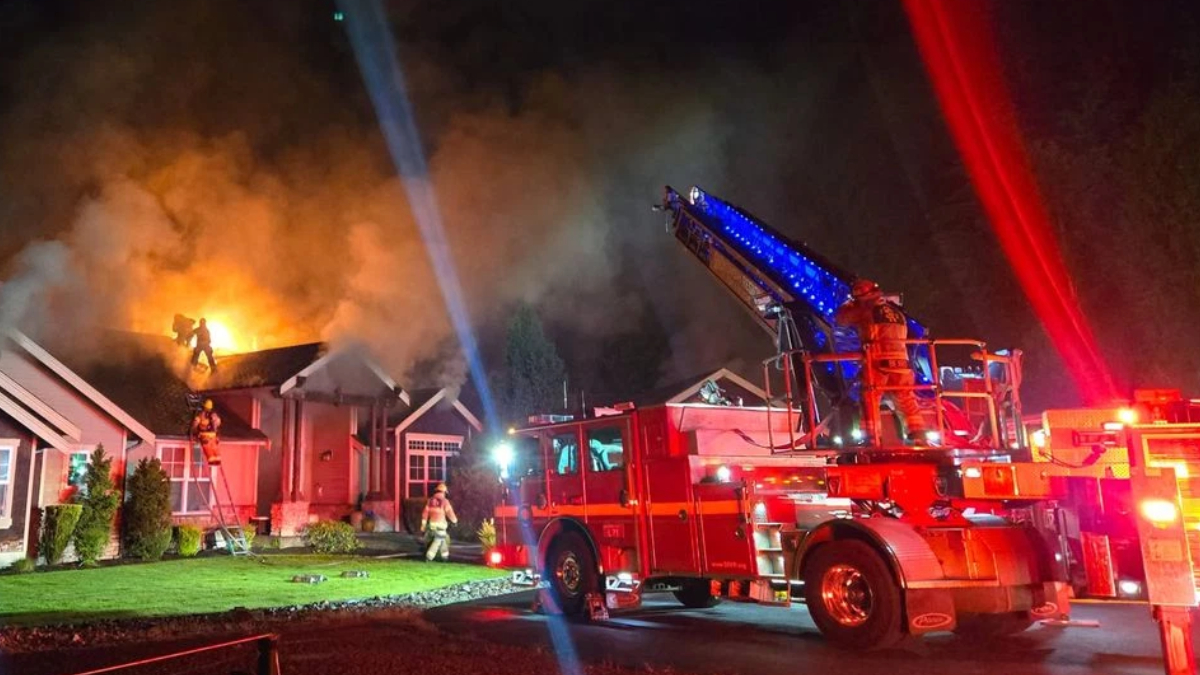 Nighttime fire at a house; flames on the roof, smoke, and firefighters on a ladder battling the blaze.