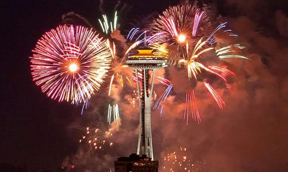 Fireworks explode around Seattle's Space Needle at night, colorful bursts lighting the sky above the city.