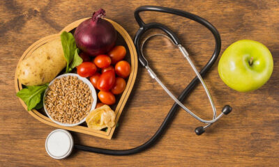Health-themed scene: a heart-shaped tray of fresh vegetables and grains on a wooden table, with a stethoscope and a green apple nearby