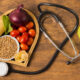 Health-themed scene: a heart-shaped tray of fresh vegetables and grains on a wooden table, with a stethoscope and a green apple nearby