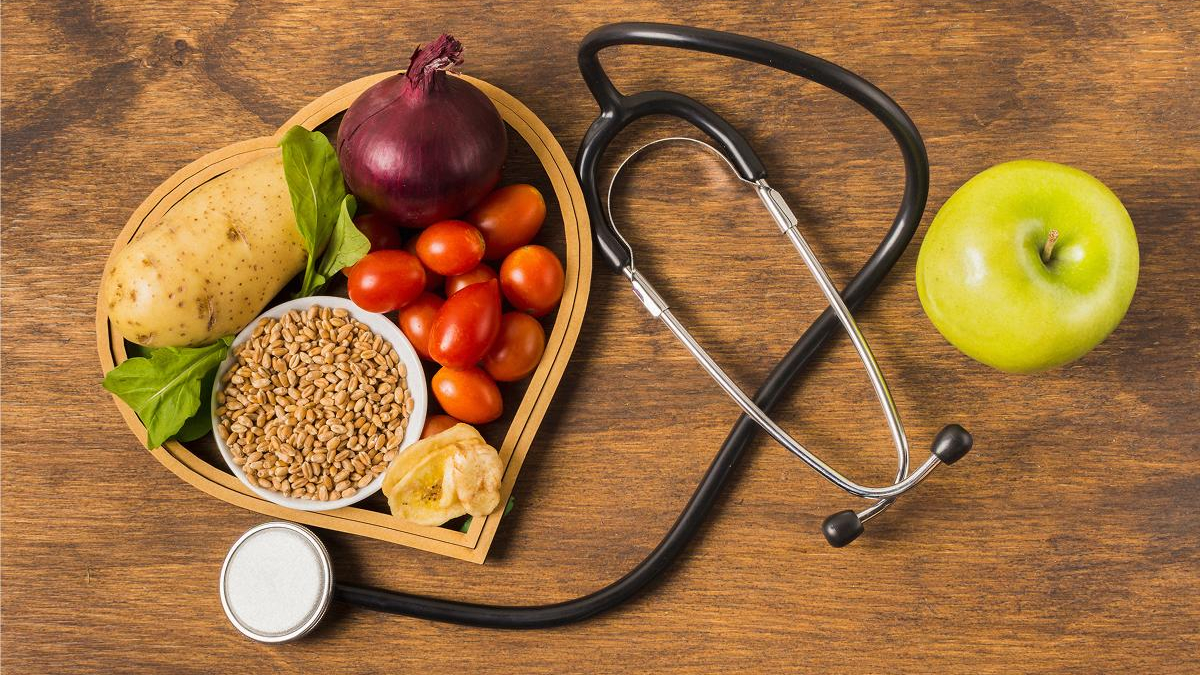 Health-themed scene: a heart-shaped tray of fresh vegetables and grains on a wooden table, with a stethoscope and a green apple nearby