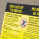 Bright yellow warning sign on a sandy beach with 'Stay Out of the Water' and Spanish translation, featuring a no-swimming symbol.