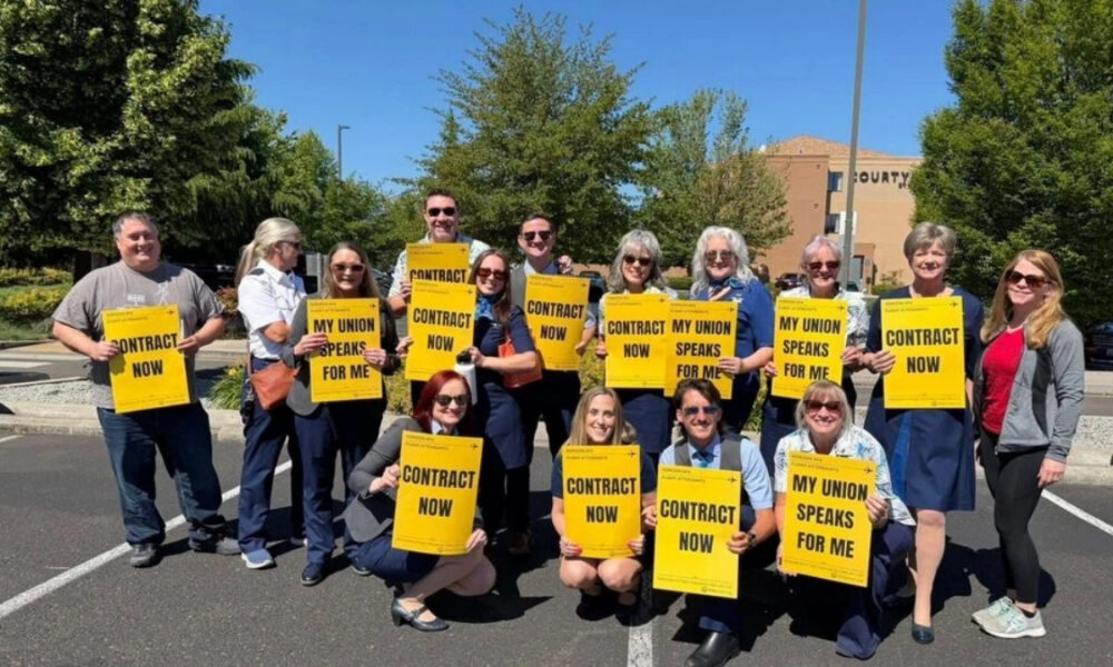 Group of adults standing in a parking lot outdoors, holding bright yellow signs that read ‘CONTRACT NOW’ and ‘MY UNION SPEAKS FOR ME,’ showing a union rally or protest.