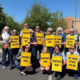 Group of adults standing in a parking lot outdoors, holding bright yellow signs that read ‘CONTRACT NOW’ and ‘MY UNION SPEAKS FOR ME,’ showing a union rally or protest.