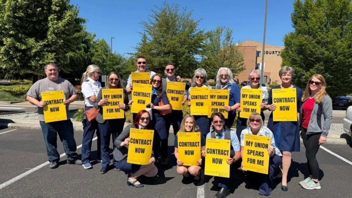 Group of adults standing in a parking lot outdoors, holding bright yellow signs that read ‘CONTRACT NOW’ and ‘MY UNION SPEAKS FOR ME,’ showing a union rally or protest.