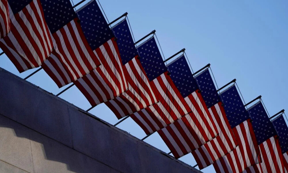 Row of American flags mounted along a railing, fluttering above a stone wall against a clear blue sky.