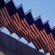 Row of American flags mounted along a railing, fluttering above a stone wall against a clear blue sky.