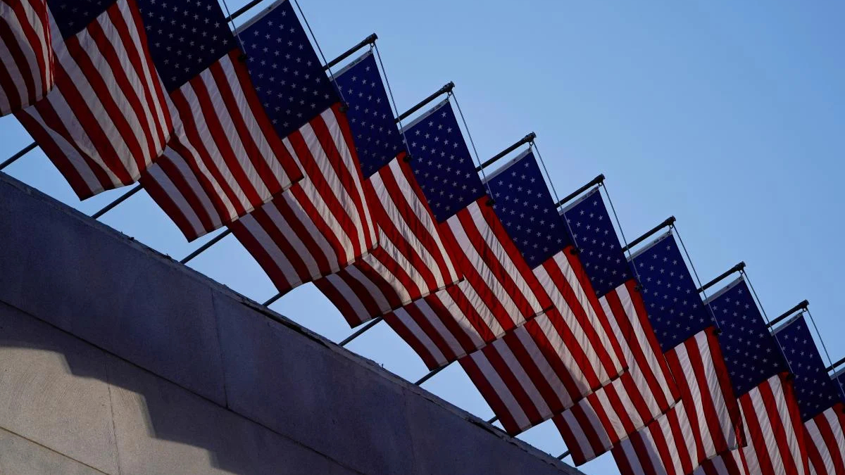 Row of American flags mounted along a railing, fluttering above a stone wall against a clear blue sky.