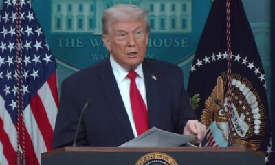 A man in a dark suit and red tie speaks at a White House podium with American flags behind him.