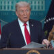 A man in a dark suit and red tie speaks at a White House podium with American flags behind him.