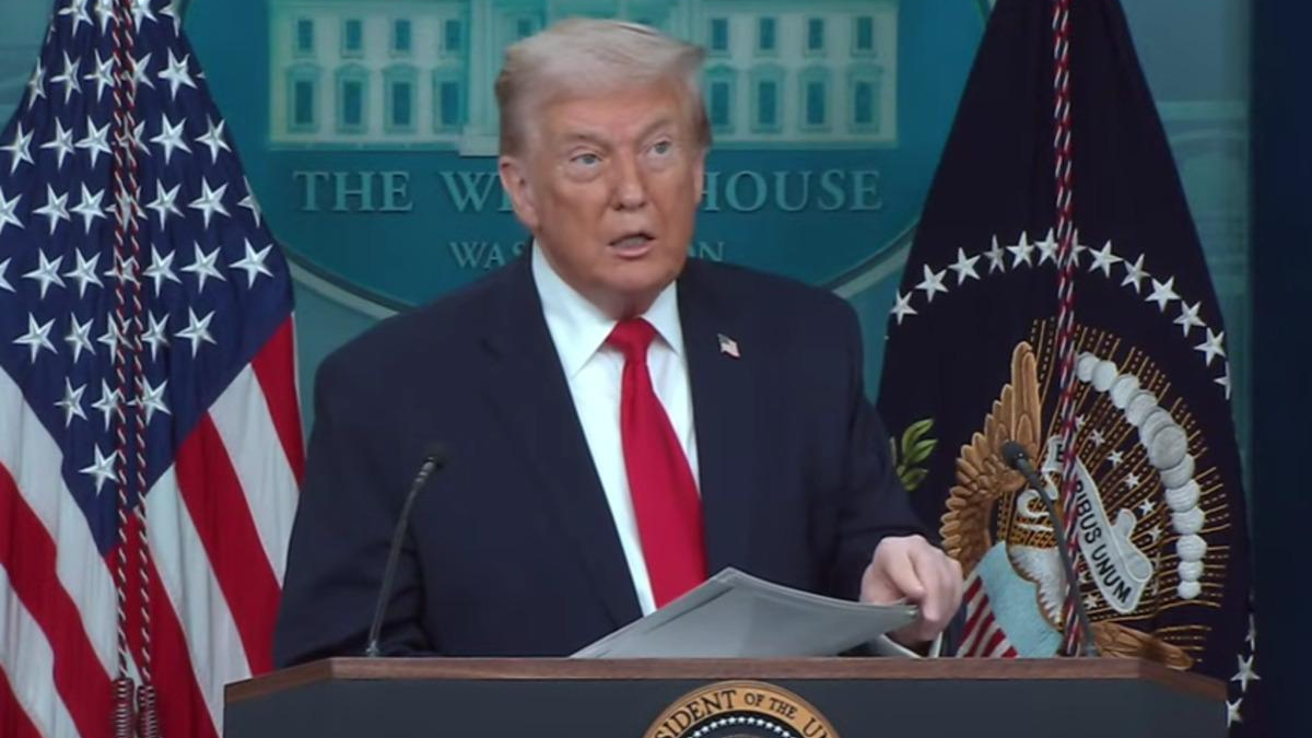 A man in a dark suit and red tie speaks at a White House podium with American flags behind him.