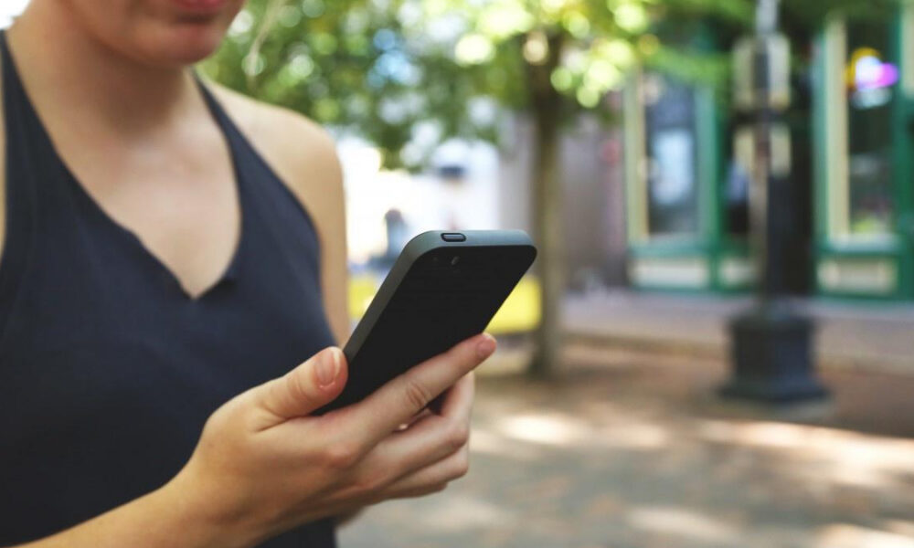 Person holding a smartphone outdoors on a sunlit, tree-lined street with blurred buildings in the background.