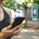 Person holding a smartphone outdoors on a sunlit, tree-lined street with blurred buildings in the background.