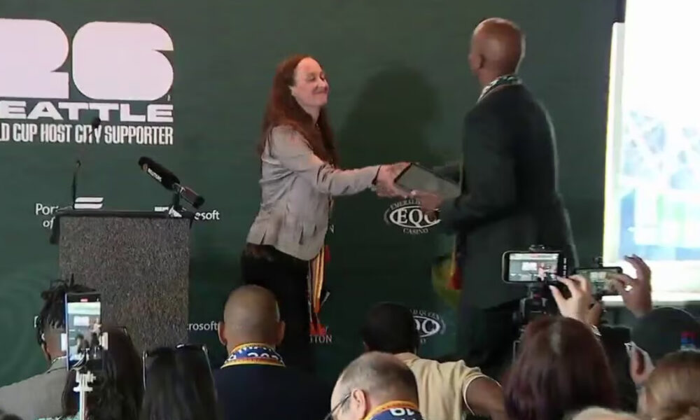 Person on stage shakes hands with a man while receiving a plaque at an event podium, audience filming nearby. Front banner reads 'SEATTLE' and event branding in green backdrop.