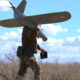 Soldier in camouflage pants carries a small unmanned aerial vehicle under his arm, holding a remote controller in a dry field.