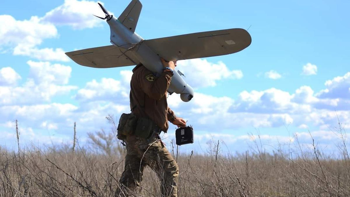 Soldier in camouflage pants carries a small unmanned aerial vehicle under his arm, holding a remote controller in a dry field.