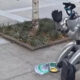 Silver humanoid robot statue on a sidewalk beside a small tree, waving with a gloved hand; a flyer and a backpack lie nearby.