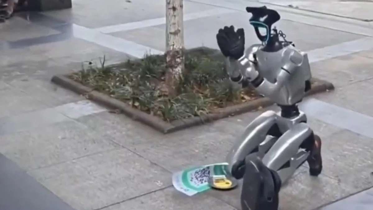 Silver humanoid robot statue on a sidewalk beside a small tree, waving with a gloved hand; a flyer and a backpack lie nearby.