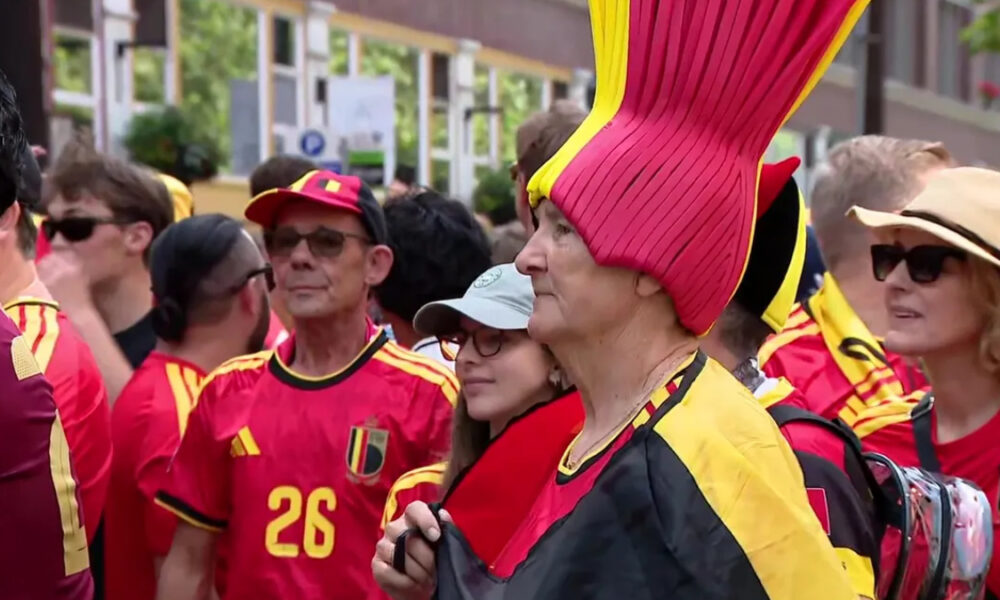 Group of Belgian football fans in red and yellow, with a woman in a tall pink-and-yellow hat in the foreground at a street event.