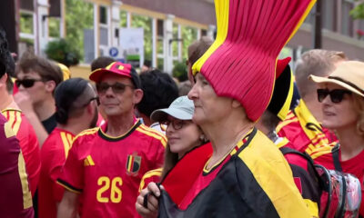 Group of Belgian football fans in red and yellow, with a woman in a tall pink-and-yellow hat in the foreground at a street event.